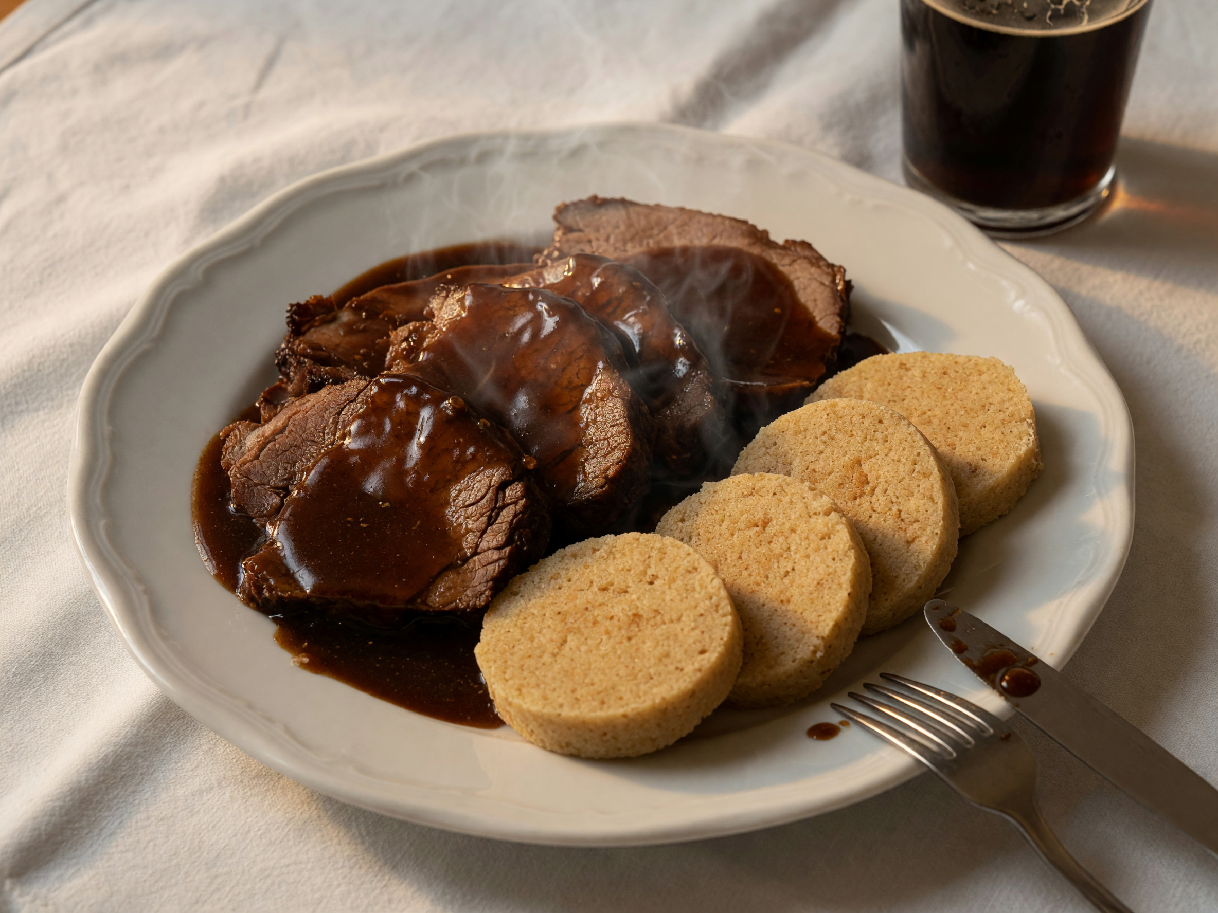 Deftiger Bratenteller mit Semmelknödeln und dunkler Sauce auf weißem Leinentischtuch.